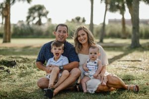 Zach Childs and his wife and two kids smiling for the camera while sitting on the grass, in the shade if the surrounding trees.
